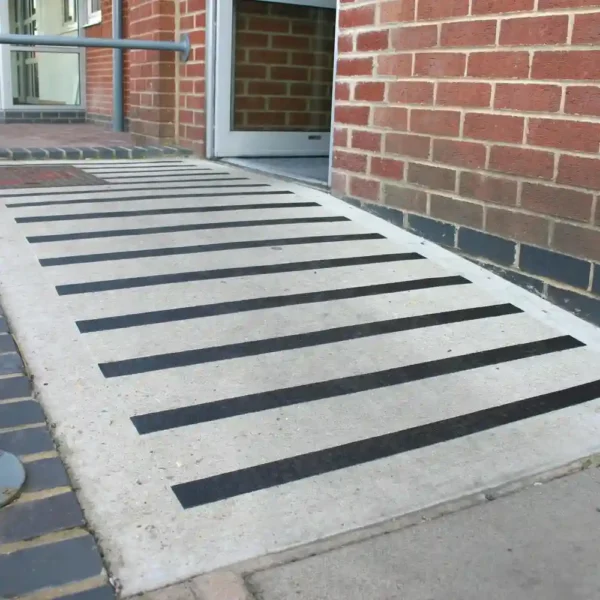A concrete wheelchair-accessible ramp with GRP Grit Tape anti-slip strips leads to a building entrance with an open door, bordered by a red brick wall and featuring a metal handrail on one side.