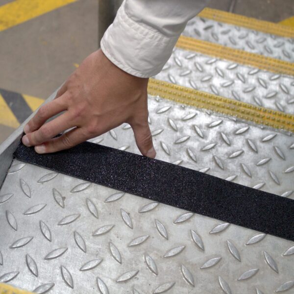 A hand applies Anti-Slip Comfortable Tape to a chequer plate metal stair tread, improving safety by adding a black anti-slip surface.