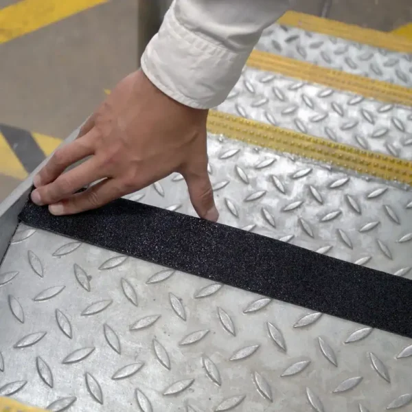 Someone applies Anti-Slip Comfortable Tape to the edge of a metal stair tread with a chequer plate pattern to increase grip and enhance safety.