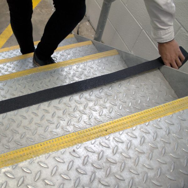 A person applies Anti-Slip Comfortable Tape, a black strip with yellow safety markings, to the edge of a metal stairway.