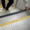 A person applies Anti-Slip Comfortable Tape, a black non-slip strip, to the edge of a metal stair with yellow safety lines in an industrial setting.