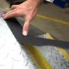 A hand applies Anti-Slip Comfortable Tape onto a metal chequer plate, with a yellow line and industrial flooring in the background.