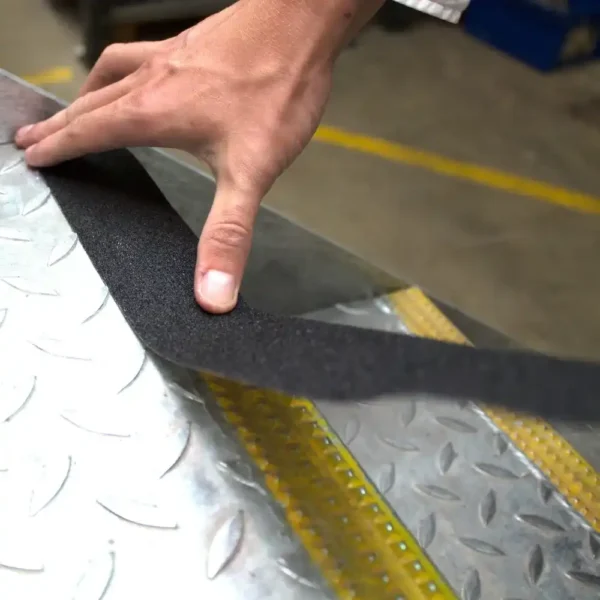 A hand applies Anti-Slip Comfortable Tape onto a metal chequer plate, with a yellow line and industrial flooring in the background.