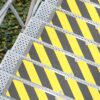An Aluminium Anti-Slip Stair Tread with perforated steps and diagonal yellow and black striped panels, beside green foliage on the ground.