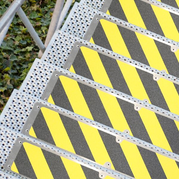 An Aluminium Anti-Slip Stair Tread with perforated steps and diagonal yellow and black striped panels, beside green foliage on the ground.