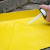 A hand uses a caulking gun to apply GRP Adhesive and Sealant onto a yellow fibreglass surface, with wooden boards in the background.
