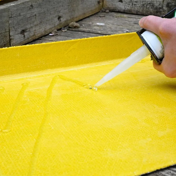 A hand uses a caulking gun to apply GRP Adhesive and Sealant onto a yellow fibreglass surface, with wooden boards in the background.