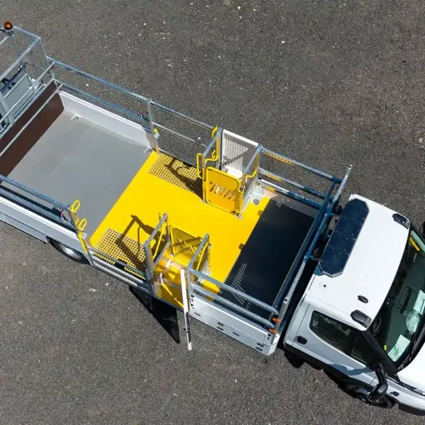 A white utility lorry with an open flatbed featuring bright yellow safety barriers and anti-slip flat sheet platforms is parked on a tarmac surface, viewed from above.