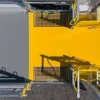 An aerial view of a bright yellow Anti-Slip Flat Sheet industrial platform with metal railings and grates, shadows on its surface, surrounded by gravel and grey flooring.