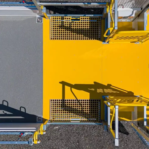 An aerial view of a bright yellow Anti-Slip Flat Sheet industrial platform with metal railings and grates, shadows on its surface, surrounded by gravel and grey flooring.