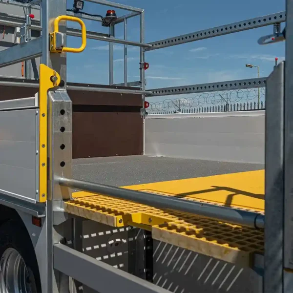 Close-up of a flatbed lorry featuring metal railings, safety barriers, and an Anti-Slip Flat Sheet yellow step platform, parked by a barbed wire fence under a clear blue sky.