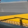 Close-up of the yellow Anti-Slip Flat Sheet on a pavement with a handrail shadow, designed for visually impaired pedestrians. A blurred grey structure and blue sky appear in the background.
