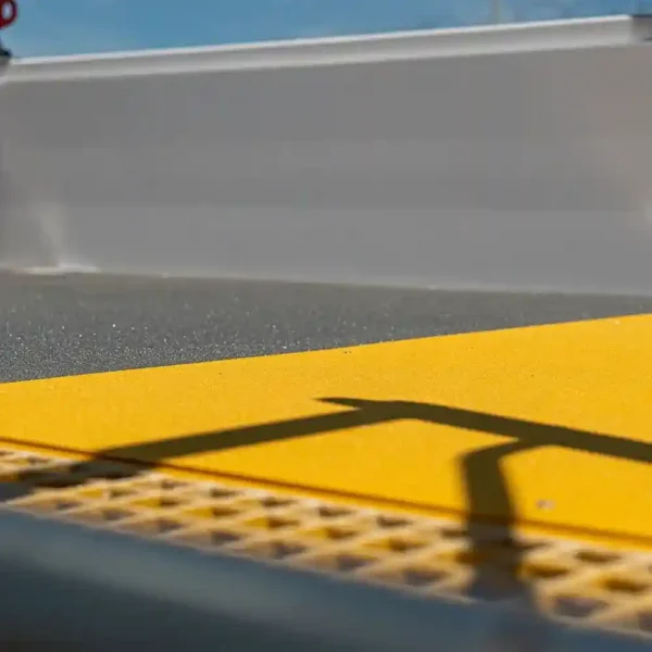 Close-up of the yellow Anti-Slip Flat Sheet on a pavement with a handrail shadow, designed for visually impaired pedestrians. A blurred grey structure and blue sky appear in the background.