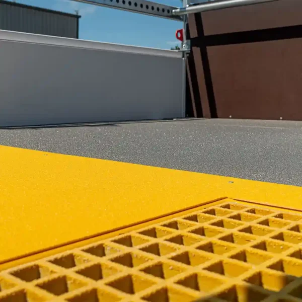A close-up of the Anti-Slip Flat Sheet shows its textured grey surface, bright yellow edge, and grated yellow section, with metal railings and a building visible in the background.