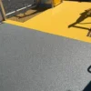 An Anti-Slip Flat Sheet in textured grey is installed beside a bright yellow section, separated by a metal barrier. The shadow of playground equipment falls across the yellow area.