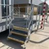 An Anti-Slip Flat Sheet covers the yellow-edged steps of a small metal staircase leading to a railed platform attached to an industrial building, near a red and white striped pole with shadows on the ground.