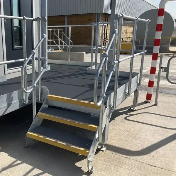 An Anti-Slip Flat Sheet covers the yellow-edged steps of a small metal staircase leading to a railed platform attached to an industrial building, near a red and white striped pole with shadows on the ground.