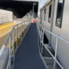 An Anti-Slip Flat Sheet with railings serves as a narrow metal walkway between a building and a yellow platform, providing safe access to a large industrial shed containing gravel or rocks.