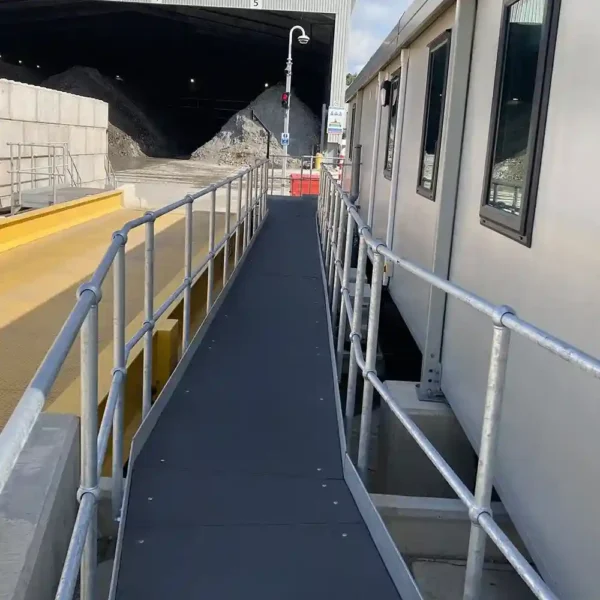 An Anti-Slip Flat Sheet with railings serves as a narrow metal walkway between a building and a yellow platform, providing safe access to a large industrial shed containing gravel or rocks.