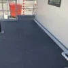A small outdoor platform features the Anti-Slip Flat Sheet flooring, with metal railings and part of a white wall with a window. In the background, there’s a red container and striped pole.