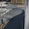 An Anti-Slip Flat Sheet in black, with yellow-striped safety edges and silver handrails, forms a wheelchair-accessible ramp leading to a building entrance beside a concrete area and wooden fence outdoors.
