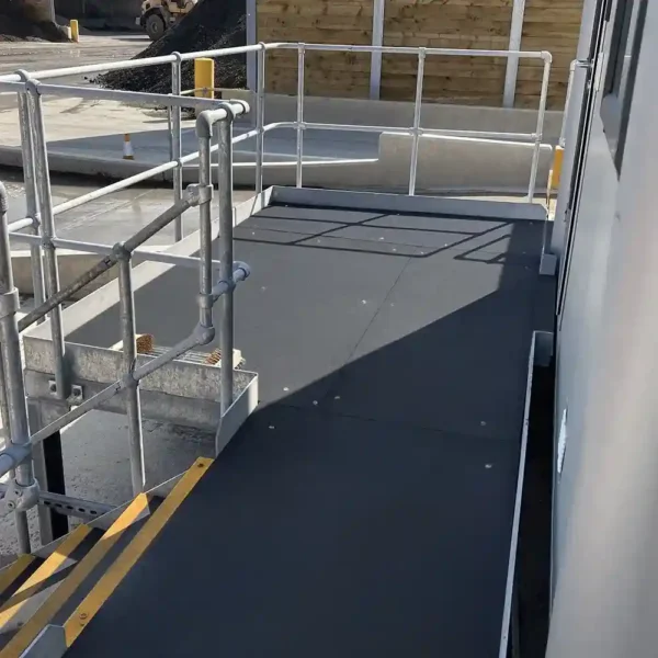 An Anti-Slip Flat Sheet in black, with yellow-striped safety edges and silver handrails, forms a wheelchair-accessible ramp leading to a building entrance beside a concrete area and wooden fence outdoors.