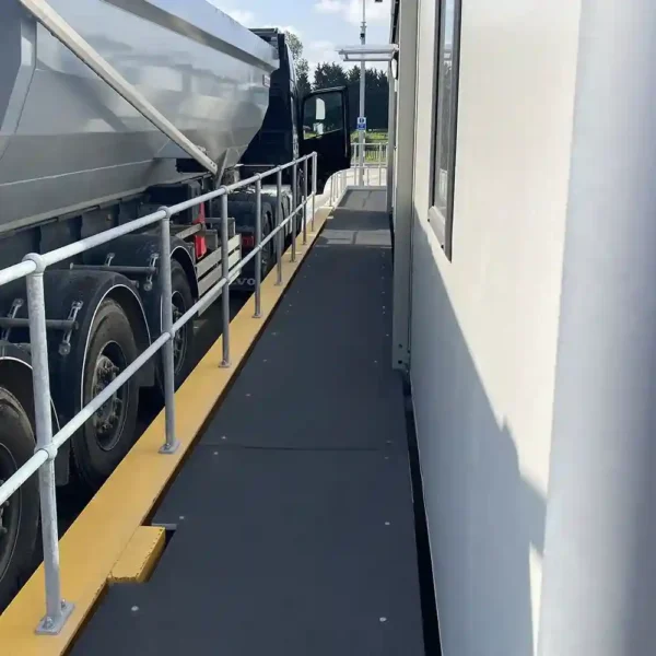 A large lorry is parked closely beside a building, separated by a narrow walkway with railings. Anti-Slip Flat Sheet is installed along the walkway edge for safety, providing secure footing between the lorry and the building.