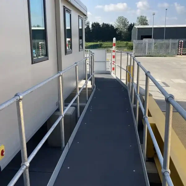 An Anti-Slip Flat Sheet covers a wheelchair-accessible ramp with metal railings, leading to a portable building in an outdoor area featuring fencing, a parked vehicle, and greenery.