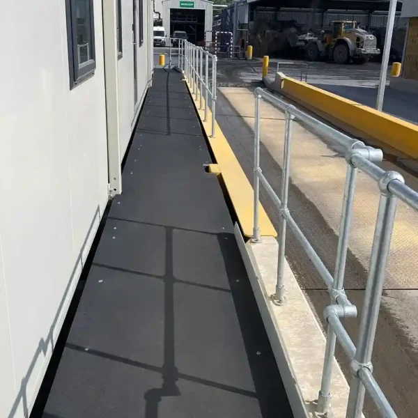A narrow walkway featuring an Anti-Slip Flat Sheet with a black non-slip surface runs beside a building, bordered by metal railings. A construction vehicle and industrial equipment are visible in the background.