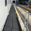 A black Anti-Slip Flat Sheet walkway with metal railings runs next to a white building, with industrial vehicles and equipment visible in the background beyond a yellow kerb.