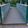 A small footbridge with metal railings features an Anti-Slip Flat Sheet as its grey surface, connecting two gravel paths and surrounded by greenery and trees.