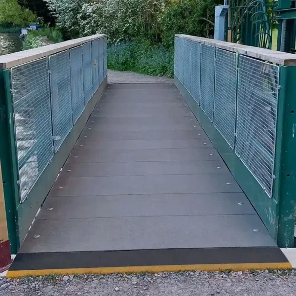 A small pedestrian bridge featuring Anti-Slip Flat Sheet decking, with green railings and metal mesh sides, spans a gravel path surrounded by greenery.