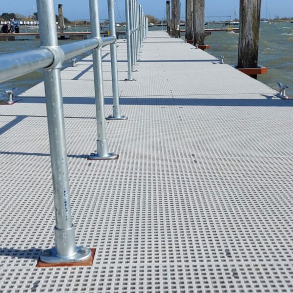 A metal-railed jetty with a Mini Mesh Grating surface extends over choppy water under a blue sky. The textured, perforated grating is visible up close, with boats and wooden posts seen in the background.