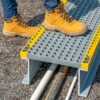 A person wearing tan work boots stands on a Step Over Platform with grey metal and yellow edges, covering cables on a gravel surface.