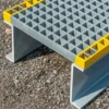 The Step Over Platform features a grey, grid-patterned metal surface with yellow edges and is elevated on sturdy angled supports at each end, positioned on rough tarmac.