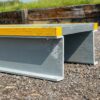A close-up of the Step Over Platform shows its grey metal beam with a yellow stripe on top, resting on gravel, while grass and wooden planks are blurred in the background.