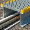 The Step-Over Platform covers several pipes and cables on a gravel surface with its gritted top and yellow edges, providing a safe crossing point.