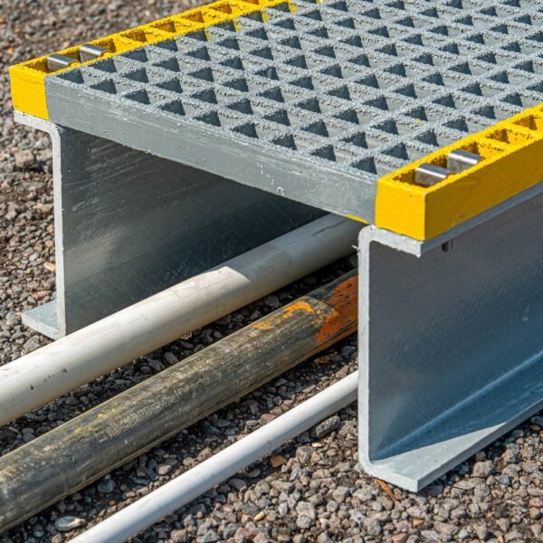 The Step-Over Platform covers several pipes and cables on a gravel surface with its gritted top and yellow edges, providing a safe crossing point.