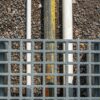 A Step-Over Platform with a metal grating and square openings is placed above three pipes—two white and one dark with a yellow stripe—resting on gravel. The top-down view highlights the textures and alignment of the pipes beneath the platform.
