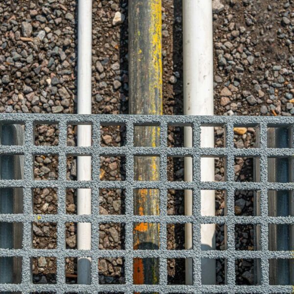 A Step-Over Platform with a metal grating and square openings is placed above three pipes—two white and one dark with a yellow stripe—resting on gravel. The top-down view highlights the textures and alignment of the pipes beneath the platform.