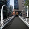 A modern pedestrian bridge with anti-slip sheets and white railings spans between glass office buildings, leading towards a red-brick building under a clear sky.