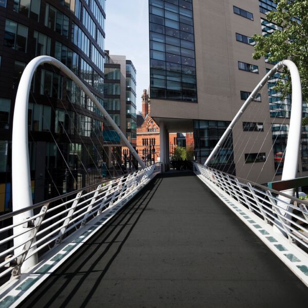 A modern pedestrian bridge with anti-slip sheets and white railings spans between glass office buildings, leading towards a red-brick building under a clear sky.