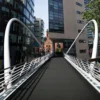 A modern pedestrian bridge with white railings and cables, equipped with the Anti-Slip Sheet, spans between office buildings and leads to a distant brick building with a clock tower.