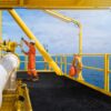 A worker in orange overalls and a white helmet operates a valve on a yellow offshore oil platform, where an Anti-Slip Sheet is attached to the railing for safety. The blue sea and sky are visible in the background.
