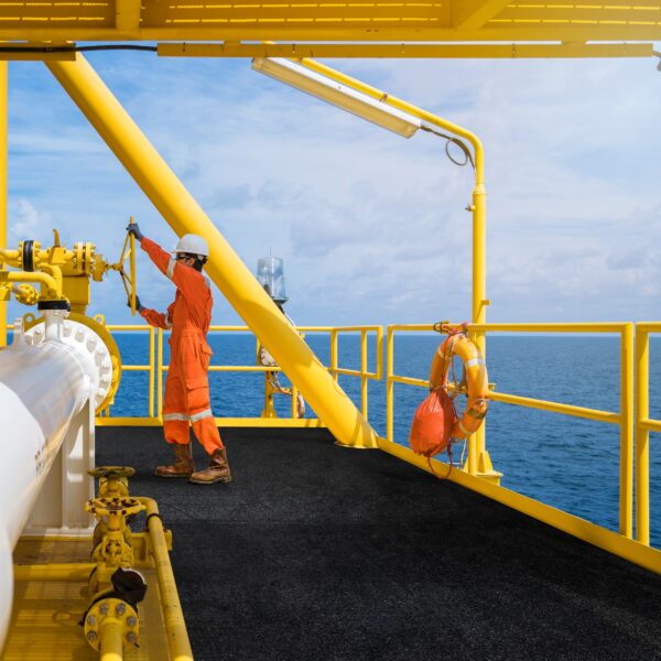 A worker in orange overalls and a white helmet operates a valve on a yellow offshore oil platform, where an Anti-Slip Sheet is attached to the railing for safety. The blue sea and sky are visible in the background.