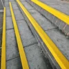 Anti-Slip Stair Nosing with yellow strips installed on concrete steps to enhance pedestrian visibility and safety, featuring a brick side wall, photographed at an angle.