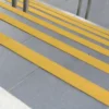 Outdoor stairs feature grey tiles and Anti-Slip Stair Nosing in bright yellow on each step for safety. Stainless steel handrails are visible at the top of the steps.