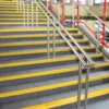 Grey stairs feature Anti-Slip Stair Nosing in bright yellow on the edges, with metal handrails in the centre and sides, red railing supports, and a backdrop of brick walls and windows.