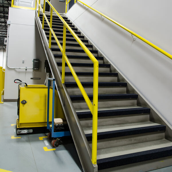 A set of black steps with Anti-Slip Stair Nosing and yellow handrails in an industrial space; a yellow trolley and blue trolley are at the base of the stairs.