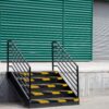 Black metal stairs with Anti-Slip Stair Nosing in yellow stripes lead to a loading bay, set before a green corrugated wall and closed grey rolling shutter door.